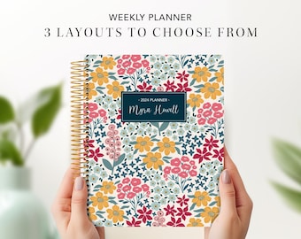 May include: A weekly planner with a floral design in shades of pink, yellow, blue, and green. The planner has a gold coil binding and a personalized name on the front cover. The text on the cover reads "2024 Planner - Myra Howell".