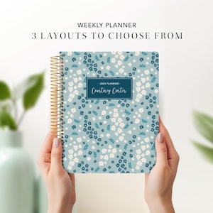 May include: A floral-patterned weekly planner with a gold spiral binding. The cover features a light blue background with white and dark blue flowers. Text on the cover reads "2024 Planner - Courtney Carter". The planner is held by two hands.