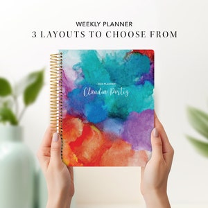May include: A colourful weekly planner with a watercolour design featuring shades of blue, purple, orange, and red. The planner has a gold spiral binding and the text "2024 Planner - Claudia Portiz" is printed on the cover.