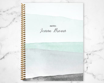 May include: A white spiral bound notebook with a watercolor design in shades of blue and gray. The cover has the text "NOTES" and "Jenna Brown" written in black.