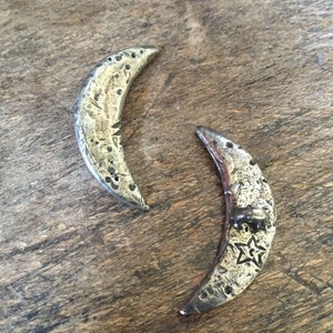 May include: Two crescent moon-shaped pendants with a textured, metallic finish. One pendant has a star design. The pendants are displayed on a wooden surface, showcasing their celestial theme.