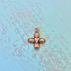 May include: A small, gold-toned metal charm in the shape of a cross with a heart in the center. The charm has a loop at the top for attaching to a necklace or bracelet.