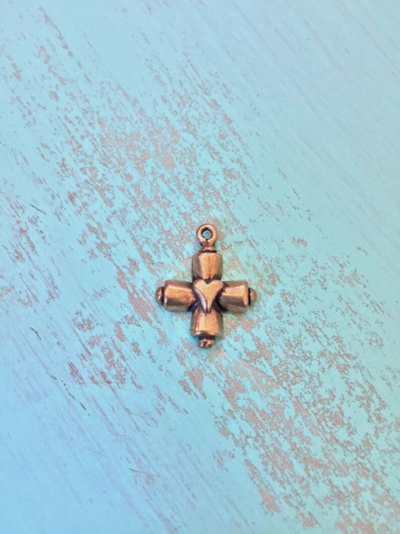 May include: A small, gold-toned metal charm in the shape of a cross with a heart in the center. The charm has a loop at the top for attaching to a necklace or bracelet.
