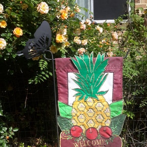 May include: A welcome garden flag with a pineapple design. The pineapple is yellow and green with red apples on the bottom. The flag is maroon with gold trim and the word "Welcome" in gold lettering.