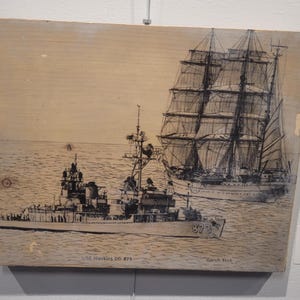 Puede incluir: Un dibujo en blanco y negro de dos barcos sobre una tabla de madera. El barco de la izquierda es un buque de guerra con el texto "USS Hawkins DD 873". El barco de la derecha es un velero. El dibujo está firmado "Gorch Fock".