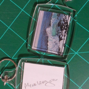 May include: Two clear acrylic keychains with silver key rings. One keychain features a print of a blue and white ocean wave. The other keychain has the handwritten text "Myke Irving 2016". Both keychains are on a green cutting mat.