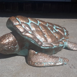 May include: A bronze-colored sea turtle statue with a teal-colored shell. The turtle is lying on its belly with its head turned to the side.