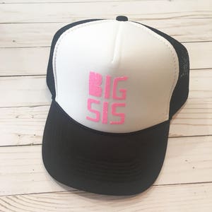 Puede incluir: Una gorra de camionero blanca y negra con un diseño de purpurina rosa que dice "BIG SIS".
