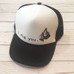 Puede incluir: Gorra de camionero blanca y negra con una malla negra en la parte posterior. La parte delantera de la gorra tiene el texto "i dig you." y un gráfico en blanco y negro de una pala.