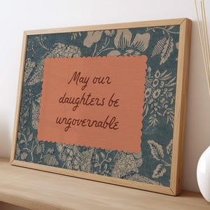 Small horizontal wall art print with "May Our Daughters be Ungovernable" in a cursive doodle font on a peach rectangle frame, layered over top of a indigo blue and cream floral print. It is in a wood frame.