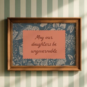 Small horizontal wall art print with "May Our Daughters be Ungovernable" in a cursive casual doodle font on a peach rectangle frame, layered over top of a indigo blue and cream floral print. It is in a wood frame.
