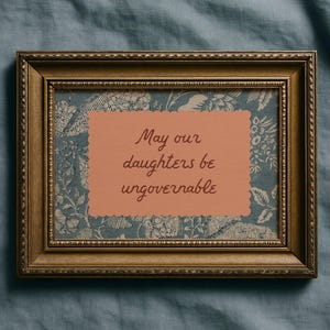 Small horizontal wall art print with "May Our Daughters be Ungovernable" in a cursive doodle font on a peach rectangle frame, layered over top of a indigo blue and cream floral print. It is in a wood frame.