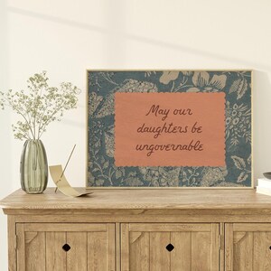 Small horizontal wall art print with "May Our Daughters be Ungovernable" in a cursive doodle font on a peach rectangle frame, layered over top of a indigo blue and cream floral print. It is in a wood frame.