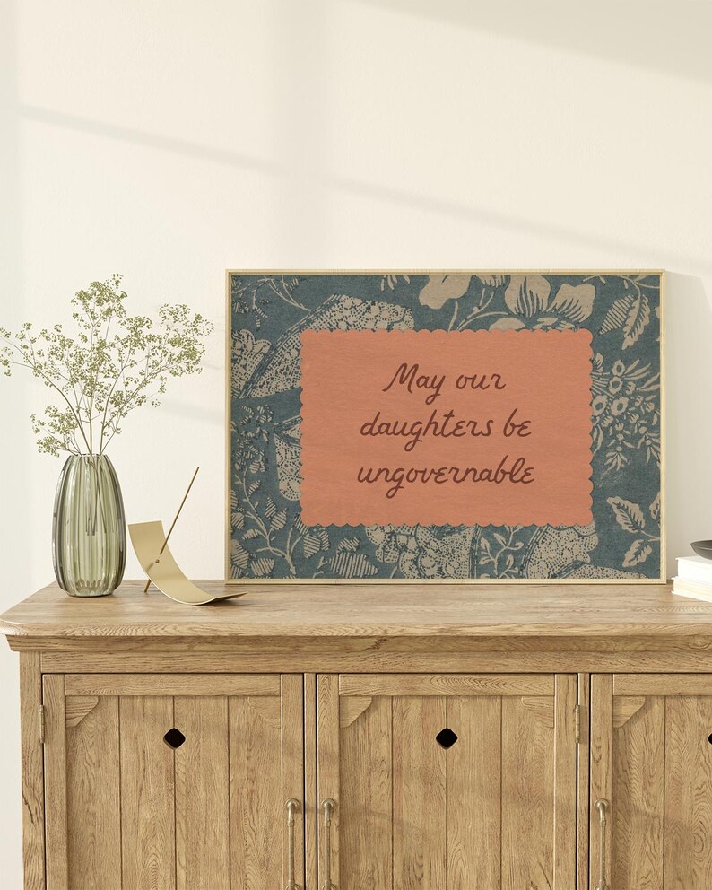 Small horizontal wall art print with "May Our Daughters be Ungovernable" in a cursive doodle font on a peach rectangle frame, layered over top of a indigo blue and cream floral print. It is in a wood frame.