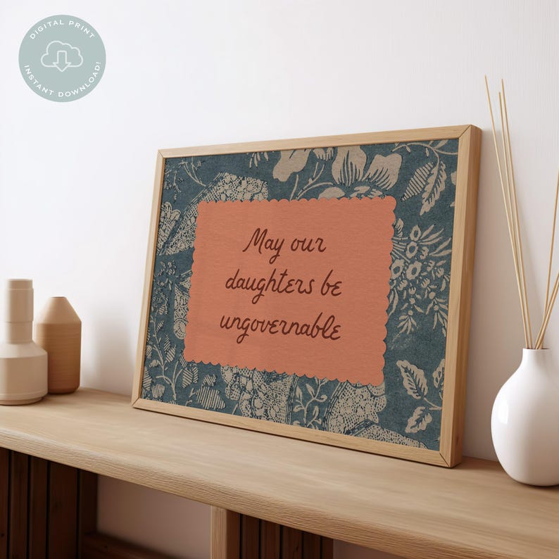 Small horizontal wall art print with "May Our Daughters be Ungovernable" in a cursive doodle font on a peach rectangle frame, layered over top of a indigo blue and cream floral print. It is in a wood frame.