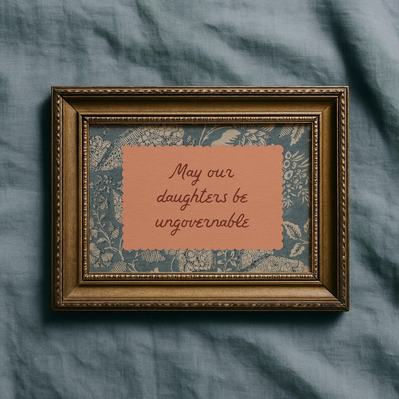 Small horizontal wall art print with "May Our Daughters be Ungovernable" in a cursive doodle font on a peach rectangle frame, layered over top of a indigo blue and cream floral print. It is in a wood frame.