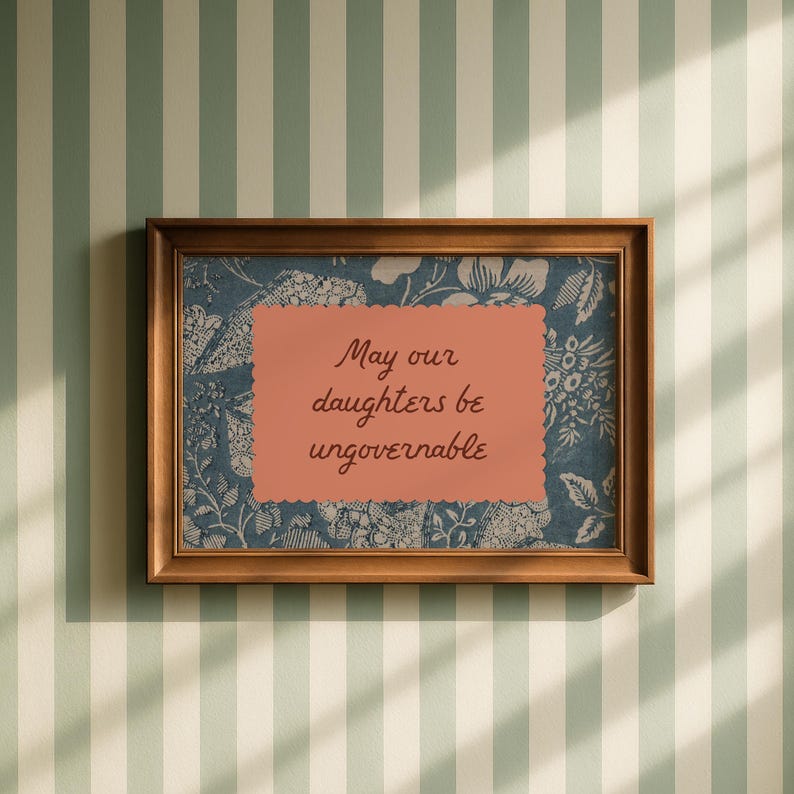 Small horizontal wall art print with "May Our Daughters be Ungovernable" in a cursive casual doodle font on a peach rectangle frame, layered over top of a indigo blue and cream floral print. It is in a wood frame.
