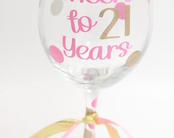 May include: A clear wine glass decorated with pink and gold polka dots and the text "Cheers to 21 Years" in pink and gold lettering. Gold and pink ribbons are tied around the stem, adding a festive touch.