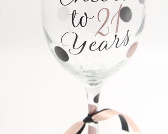 May include: A clear glass wine glass decorated with black and rose gold polka dots. The text "Cheers to 21 Years" is printed in black and rose gold script on the front. A black and rose gold ribbon is tied around the stem of the glass.