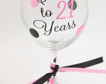 May include: A clear wine glass decorated with pink and black polka dots and the text "Cheers to 21 Years" in black and pink. The base of the glass is adorned with a pink and black ribbon.