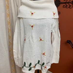 May include: A white, long-sleeved sweater with a cowl neck. The sweater is decorated with embroidered holly and berry sprigs in gold, red, and green. Small, gold-colored beads are scattered across the front. The bottom hem features a green holly garland.