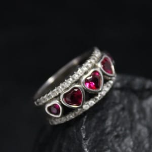 May include: A silver ring with five red heart-shaped gemstones and a row of small clear gemstones.
