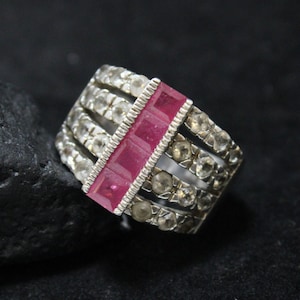 May include: A silver ring with a row of pink rectangular stones set in the center. The ring is surrounded by smaller clear stones.
