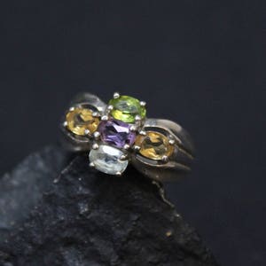 May include: A silver ring with four gemstones: a green, a purple, and two yellow. The ring is on a dark gray rock.