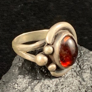 May include: A silver ring with a red gemstone set in a bezel setting. The ring has a unique design with three small silver balls on the band.