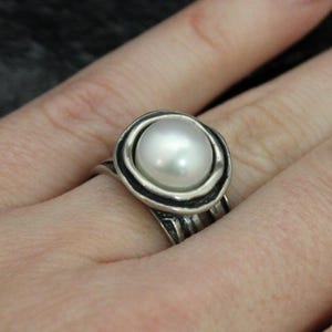 May include: A silver ring featuring a large, round, white pearl. The pearl is set in a decorative bezel with a textured band. The ring is shown on a hand with long, manicured nails.