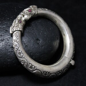 May include: A silver bangle bracelet with intricate floral designs and two red gemstones. The bracelet is hinged and opens for easy wear.