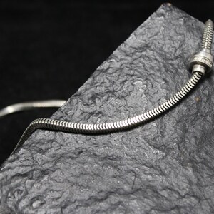 May include: A silver snake chain necklace with a silver clasp. The necklace is lying on a dark gray textured surface.