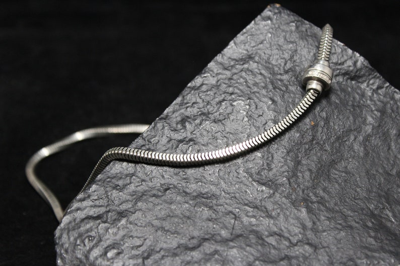 May include: A silver snake chain necklace with a silver clasp. The necklace is lying on a dark gray textured surface.