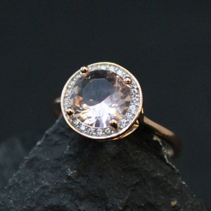 May include: A rose gold ring with a large, round, pale pink gemstone surrounded by a halo of smaller, clear gemstones.