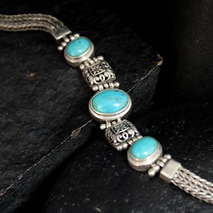 May include: Silver bracelet featuring oval turquoise stones set in ornate bezels. The bracelet has a woven chain design and a secure clasp. A beautiful piece of jewelry.