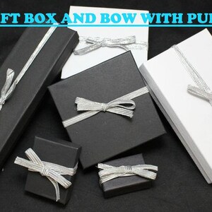 May include: Five gift boxes with silver ribbon bows. Two boxes are black and three are white. The boxes are arranged on a black surface. The text "FREE GIFT BOX AND BOW WITH PURCHASE" is at the top of the image.