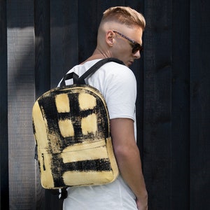 May include: A beige backpack with black paint splatter design. The backpack has a zippered pocket on the front and adjustable straps.