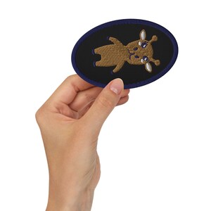 May include: A brown embroidered patch with a cartoon deer design on a black background. The patch is surrounded by a dark blue border.