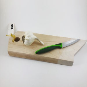 Könnte beinhalten: Ein Schneidebrett aus Holz mit einer hellen Holzoptik und einem Loch in der Mitte. Eine Knoblauchzehe und ein grünes und silbernes Küchenmesser liegen auf dem Brett.