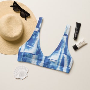 Peut inclure: Un haut de bikini bleu et blanc tie-dye avec un décolleté en scoop. Le haut est posé à plat sur une surface blanche avec un chapeau de paille, des lunettes de soleil et de la crème solaire.