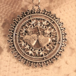 May include: Silver-toned pendant necklace featuring a detailed circular design. The pendant showcases two hearts, a cross, and floral accents within a decorative border. The necklace hangs on a delicate chain, suitable for everyday wear.