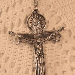 Könnte beinhalten: Silberfarbener Anhänger mit einer Halskette, die ein Kruzifix mit einer Darstellung von Jesus Christus am Kreuz zeigt. Das Kruzifix ist mit einem kreisförmigen Bild von Gott dem Vater gekrönt.