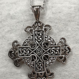 May include: Silver-toned Celtic cross pendant on a silver chain. The pendant features intricate knotwork and a circular design at the center. The cross is suspended against a light gray fabric background.