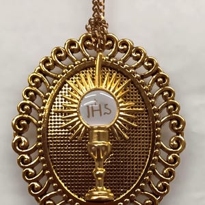 Puede incluir: Collar con colgante dorado con un marco ovalado adornado y un diseño de rayos de sol. El centro tiene un círculo blanco con las letras IHS. El colgante cuelga de una cadena dorada a juego.