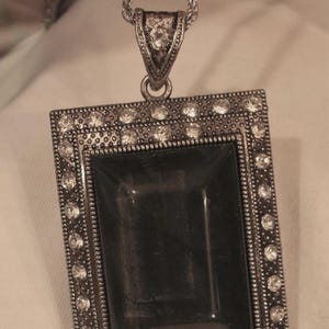 May include: A silver chain necklace with a square pendant featuring a black stone surrounded by clear rhinestones.