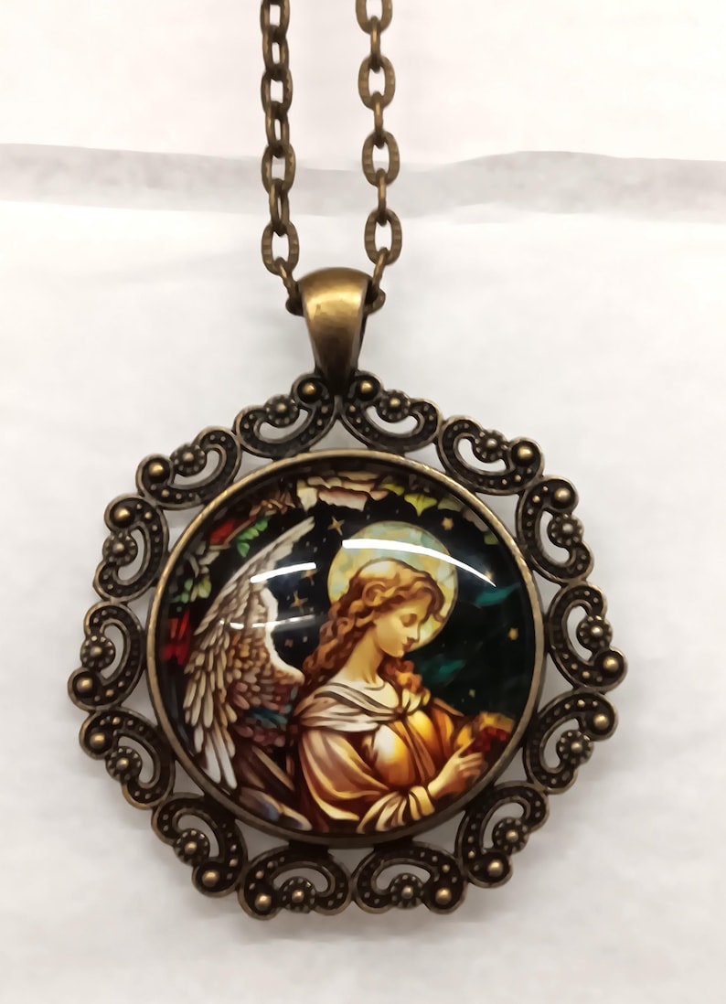 Puede incluir: Collar con colgante de tono bronce antiguo con una imagen redonda de estilo vidriera de un &aacute;ngel. El colgante est&aacute; engastado en un marco ornamentado de estilo filigrana y cuelga de una cadena a juego.