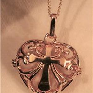 May include: Rose gold heart-shaped pendant with an ornate filigree design and a cross in the center. The pendant hangs from a delicate chain. The background is a soft, neutral color.