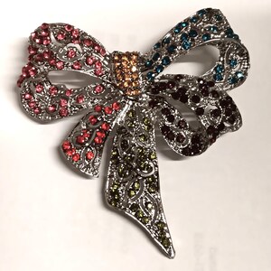 May include: A silver bow brooch embellished with a variety of coloured rhinestones. The bow features red, pink, blue, green, and brown stones, creating a sparkling accessory. The brooch has an intricate, openwork design.