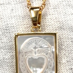 Op de afbeelding: Een gouden ketting met een hanger met een wit hart met een duifontwerp in een gouden frame.