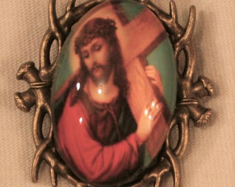 Op de afbeelding: Een bronzen broche met een ronde glazen cameo met een afbeelding van Jezus die een kruis draagt. De cameo is omgeven door een ontwerp van een doornenkroon.